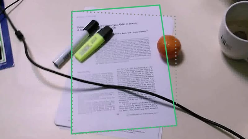 A page on a cluttered desk under markers and a cable; the model's dotted grey corners take in the sheet beneath, the true outline is green.